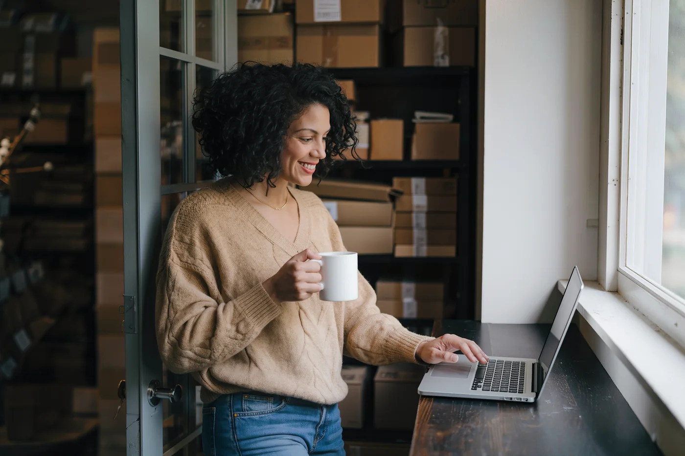 A webshop owner with a coffee, relaxed at their laptop in the stockroom